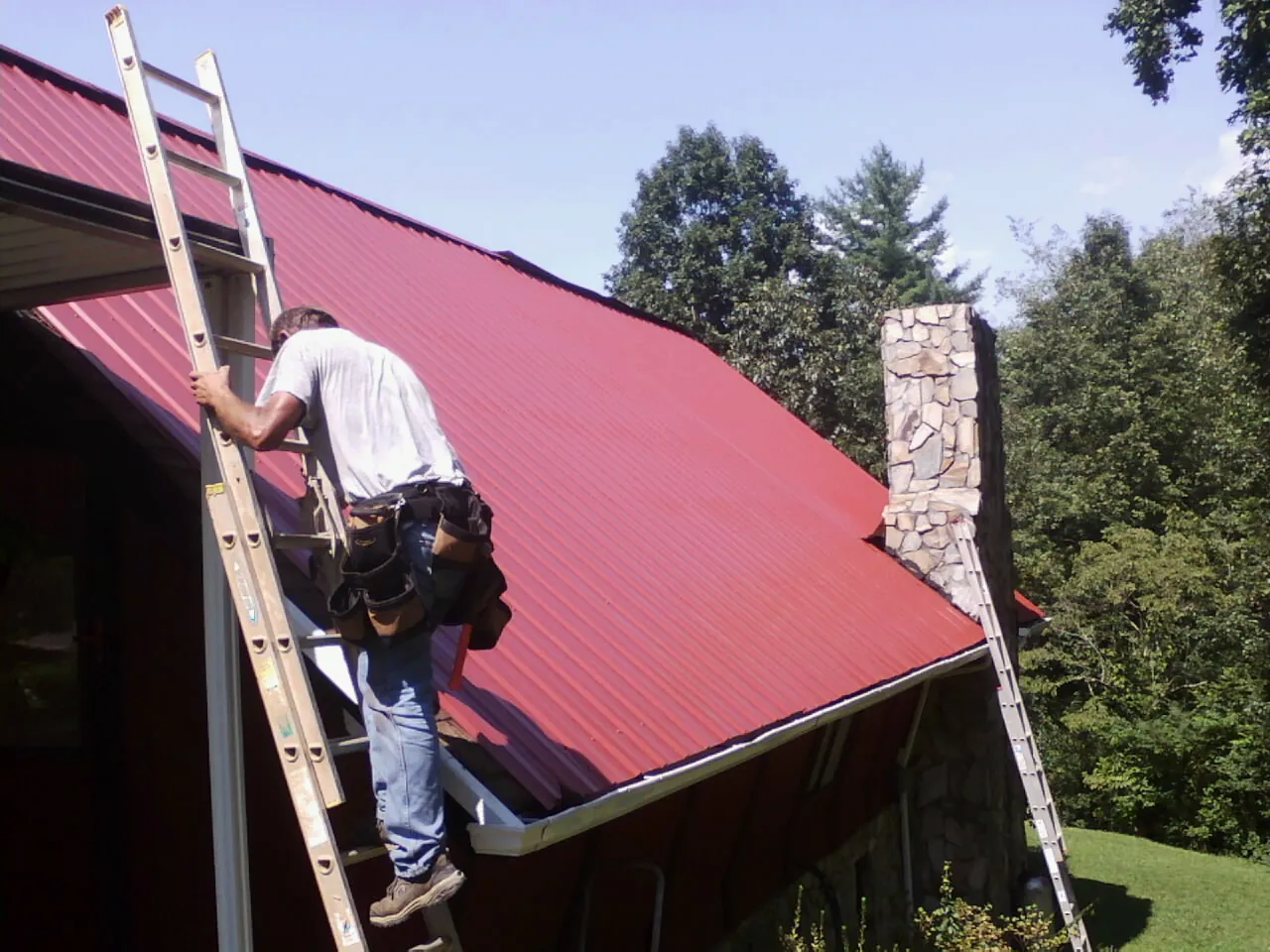 Roofer on a ladder beside a red metal roof and stone chimney — Soffit & Fascia Repair by Roofing Brothers in Redwood